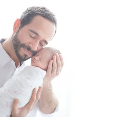 A father gently cradles his baby boy, both with eyes closed, sharing a peaceful and loving moment against a bright, white background.