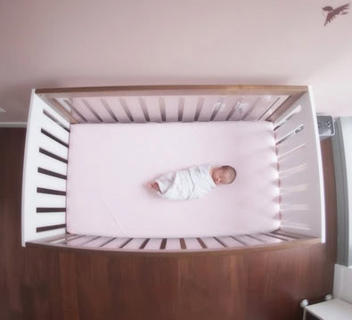 A newborn baby, swaddled in a white blanket, lies in the center of a crib with a pale pink mattress. The crib has white slats and is placed on a dark wooden floor.