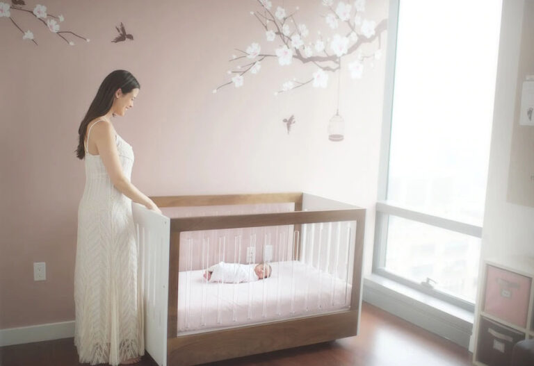 A mother in a white dress stands beside a wooden crib, looking at her swaddled infant. The nursery in the apartment features light pink walls with blossom and bird decals, and a large window bathing the room in natural light.