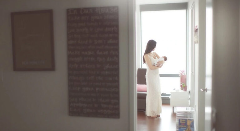A woman in a white dress stands in a bright room, gently holding a baby. Soft light streams through large windows, creating a calm, serene atmosphere. The scene is viewed from an adjacent hallway.
