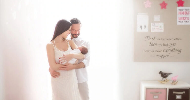 A couple stands together in a softly lit room, smiling as they hold their newborn baby wrapped in a white blanket—a heartwarming family photoshoot moment. The background features a wall with inspirational quotes and decorative items.