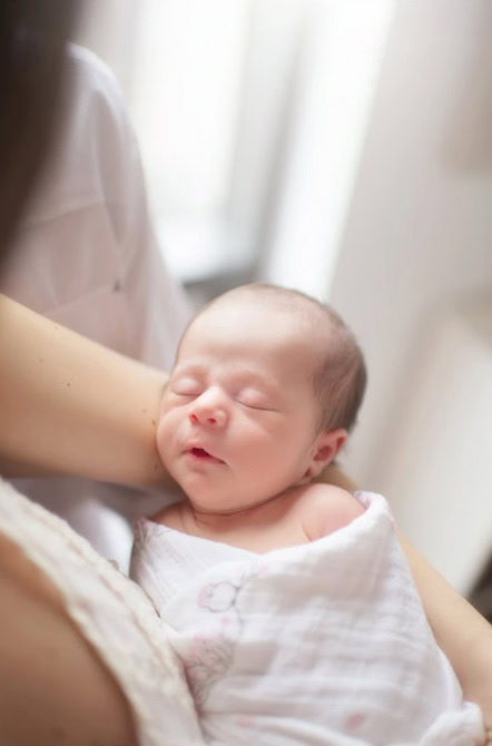 A newborn baby wrapped in a white blanket sleeps peacefully in someone's arms, bathed in soft, natural light.