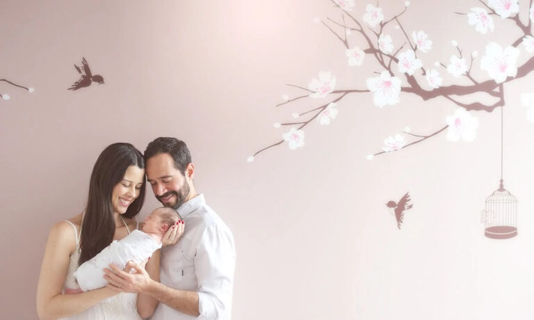 A smiling mother and father gently hold their infant baby, standing in front of a pale pink wall adorned with cherry blossom branches, birds, and an empty birdcage.