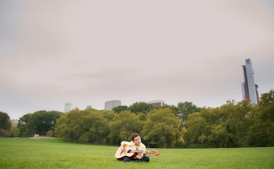 A child sits on green grass in Sheep Meadow, Central Park, playing an acoustic guitar. Trees and tall city buildings rise in the background under a cloudy sky.