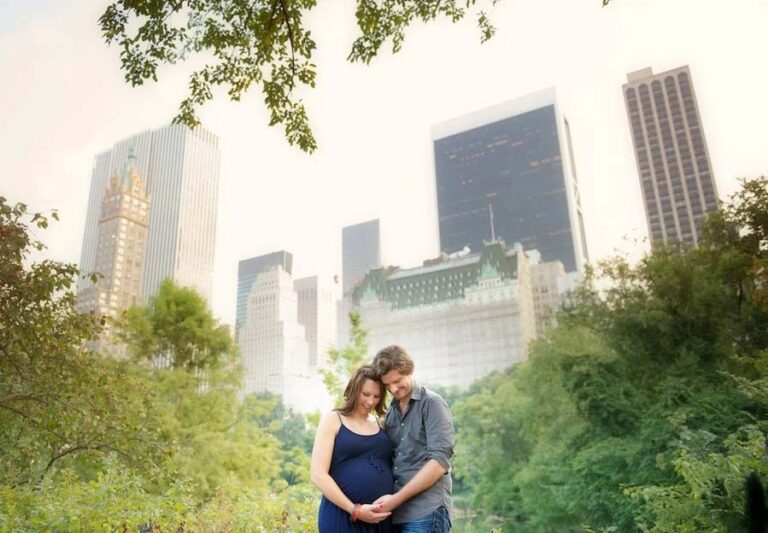 A pregnant woman in a blue dress and a man stand close together, smiling and touching her belly during a maternity photoshoot, surrounded by greenery with tall city skyscrapers in the background.