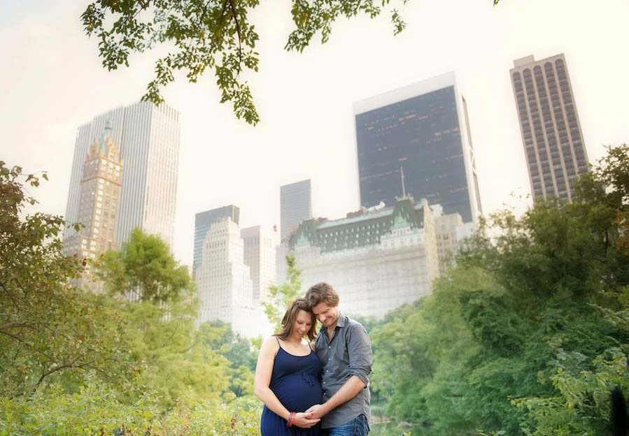 A pregnant woman in a blue dress and a man stand close together, smiling and touching her belly during a maternity photoshoot, surrounded by greenery with tall city skyscrapers in the background.