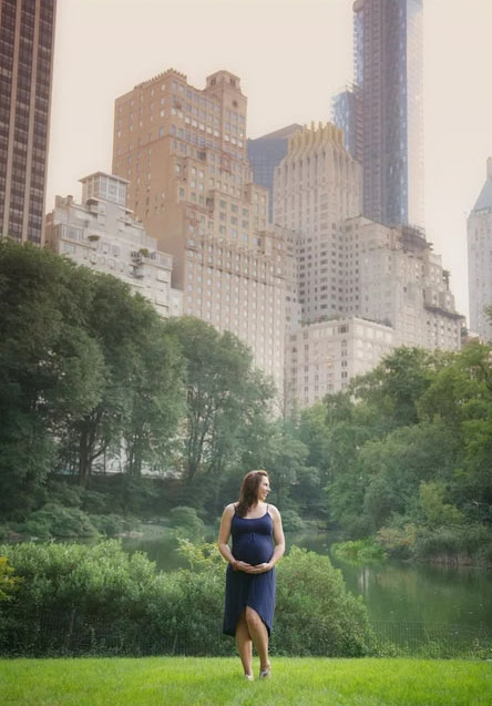 A woman in a blue dress stands on grass in Central Park, cradling her pregnant belly, with tall city buildings and lush green trees in the background.