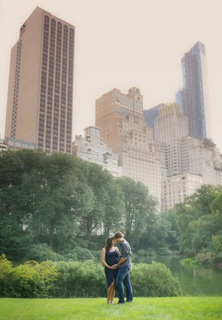 A couple stands together in a grassy park, embracing and touching the pregnant lady's belly, with lush green trees and the NYC skyline rising in the background.