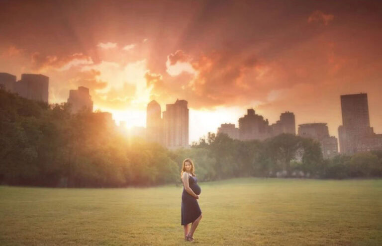 A pregnant woman in a dark dress stands in a sunlit field, city skyline behind her, as sunset Central Park maternity photographer captures warm rays streaming through dramatic clouds for a glowing atmosphere.