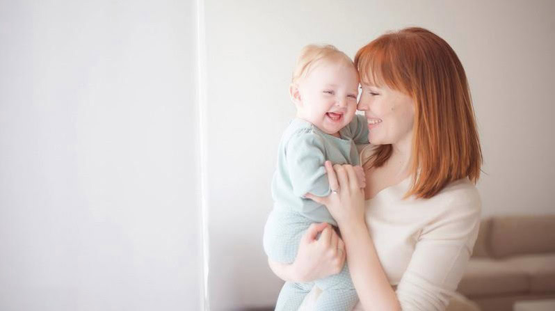A mother with red hair holds her smiling daughter with light hair, both looking happy and close together in a softly lit, cozy modern NYC apartment.