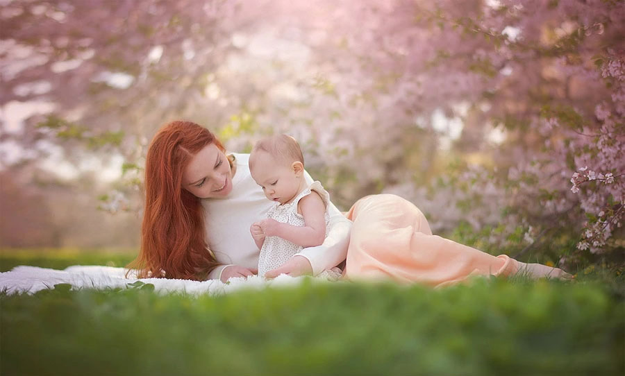 A mother and daughter with long red hair sit together on a blanket outdoors, surrounded by blooming pink flowers and greenery. The mother smiles warmly at her daughter, who is focused on something in her hands.