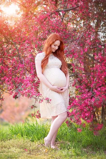 A pregnant woman with long red hair in a light dress stands barefoot on grass, gently cradling her belly amid vibrant pink flowering branches—a serene moment captured in true NYC maternity photography style.