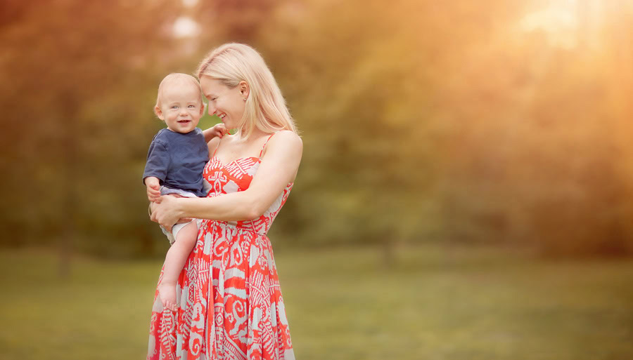 A woman in a red patterned dress holds a smiling baby outdoors, with soft sunlight and blurred greenery in the background. Captured by a NYC family photographer, both appear happy and relaxed.