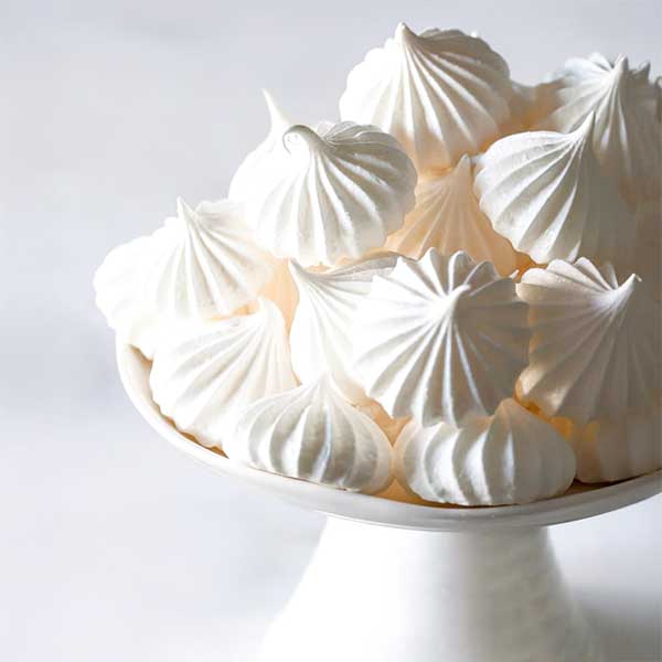A white pedestal dish filled with delicate, white meringue cookies, each with a swirled, ridged texture and a crisp appearance, against a light background.