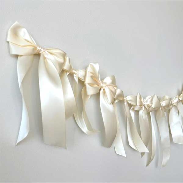 A garland of large, ivory satin bows with long trailing ribbons hangs against a plain white wall.