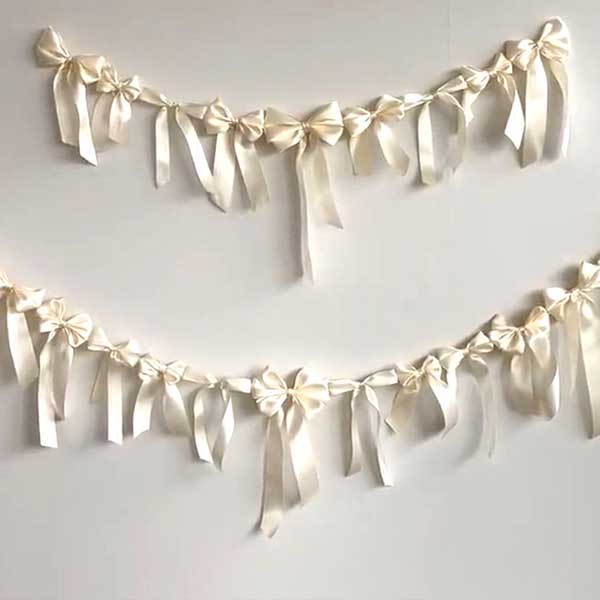 Two garlands made of cream-colored satin ribbon bows are hanging against a plain white wall. Each garland features several bows spaced evenly along the string.