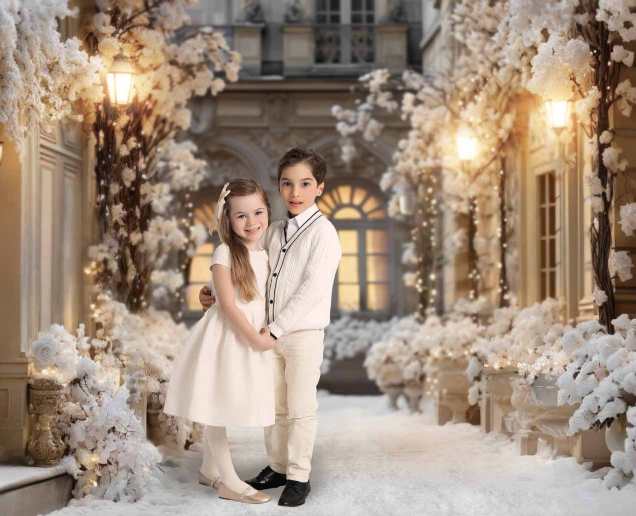 Two children in white dressy clothes stand close together, holding hands and smiling in a snowy, elegant courtyard at an NYC photo studio, surrounded by white flowers and glowing lamps that evoke a festive, winter atmosphere.