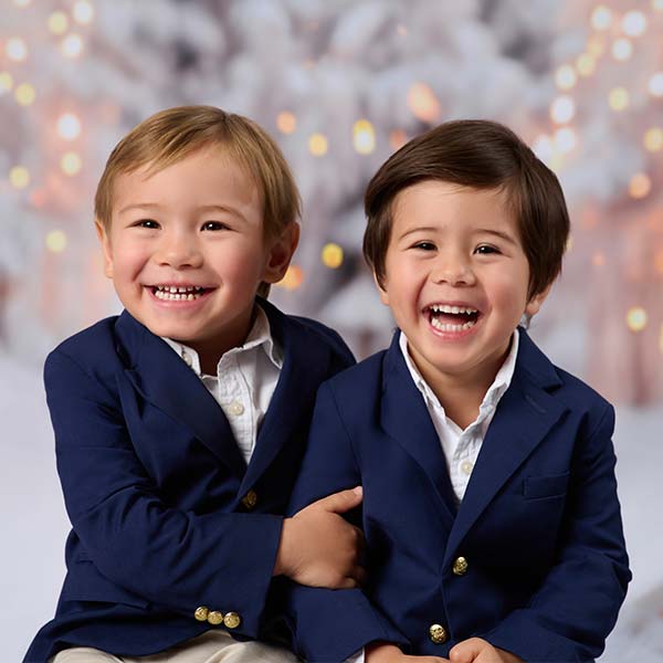 Two young boys in navy blazers and white shirts sit close together, smiling and laughing in front of a soft, blurred background with warm lights—a perfect moment captured by a family photographer in a festive or holiday setting.