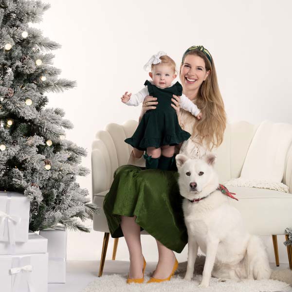 A smiling woman sits on a white couch holding a baby in a green dress and bow. A white dog with a festive bandana sits by her, next to a decorated Christmas tree and white gift boxes.