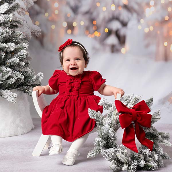 A smiling baby in a red dress and white tights sits on a white chair next to a frosted Christmas tree with a red bow. The background features snowy trees and warm, festive lights.