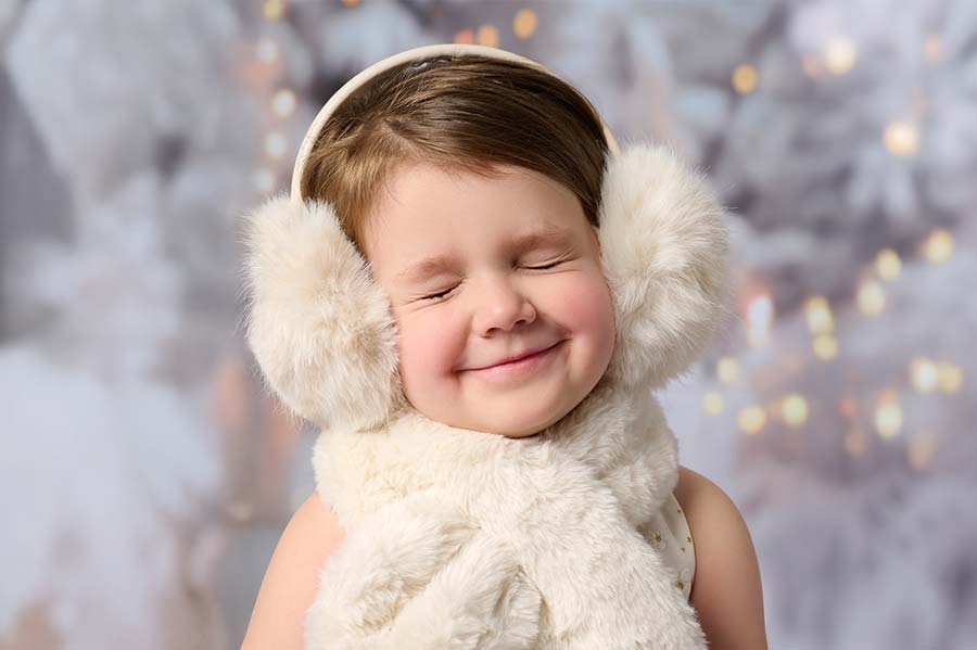 A young child smiles with eyes closed, wearing fluffy white earmuffs and a matching scarf, against a soft, snowy background with twinkling lights.