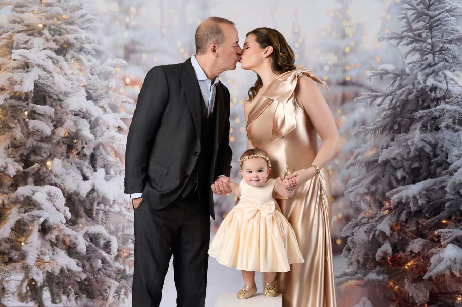 A couple dressed formally kisses while holding hands with a smiling toddler in a cream dress. Captured at an NYC photography studio, they stand before snowy, decorated trees adorned with warm lights.
