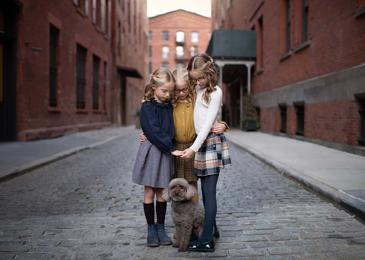 Three young girls with similar wavy blonde hair, wearing skirts and sweaters, stand closely together on a cobblestone street, gently touching a small, curly brown dog in front of them. Red brick buildings line the street.