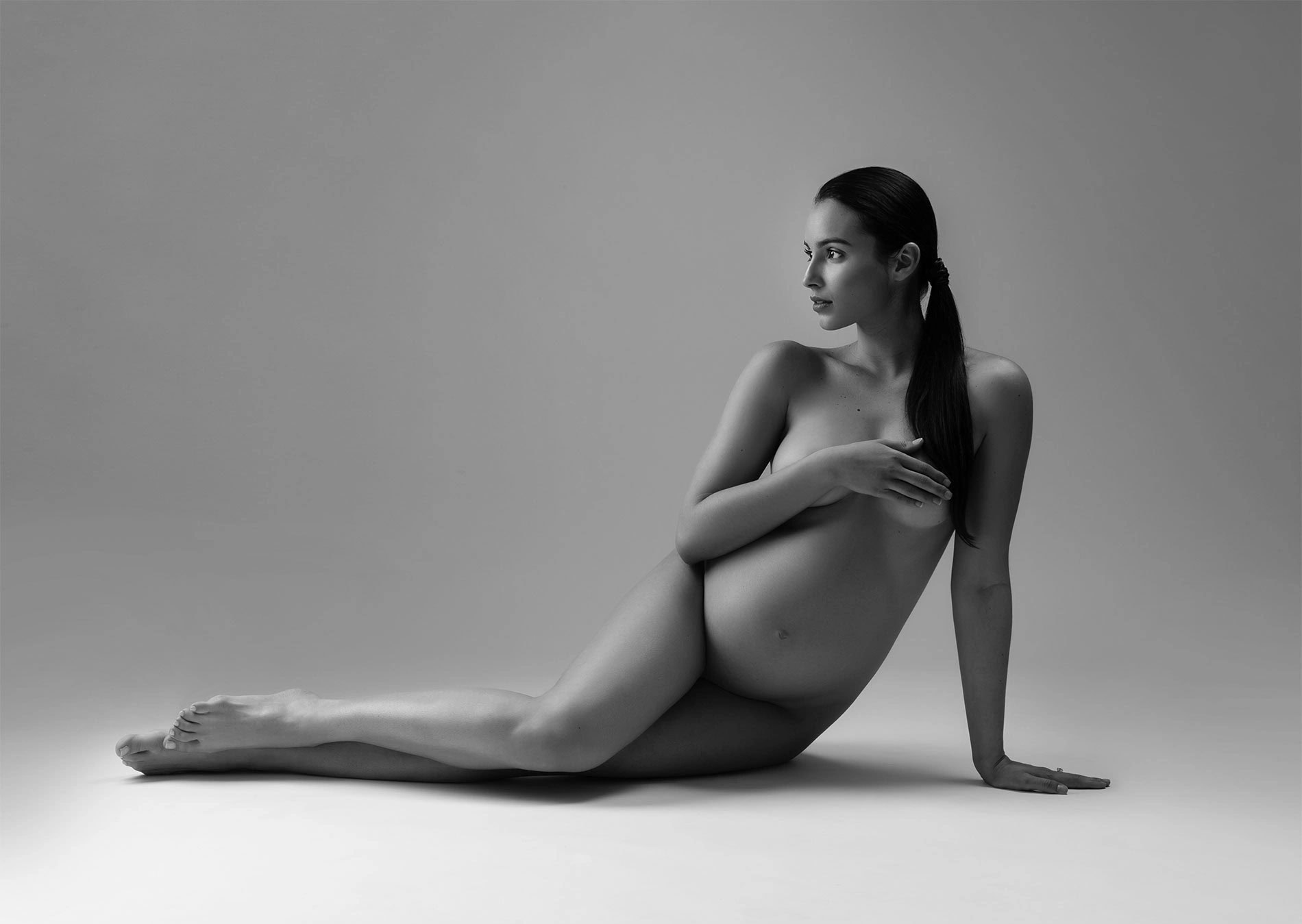 A pregnant woman poses nude in a studio, sitting on the floor with legs extended and one arm supporting her while the other covers her chest. The image is in black and white with soft lighting.