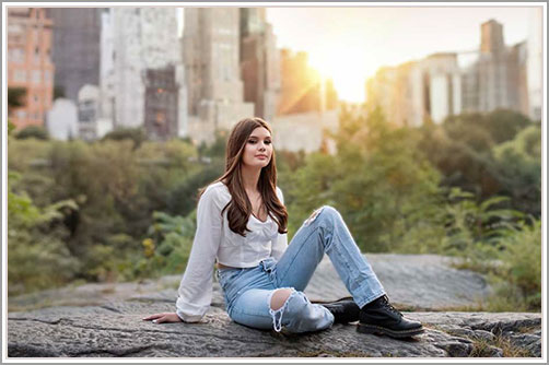 High school senior posing in Central Park, with NYC's iconic skyline in the background. The sun is setting behind the buildings, casting a warm glow over this beautiful portrait.