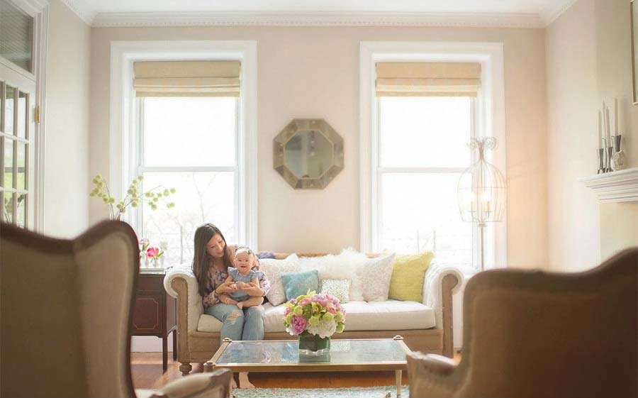 A woman sits on a beige sofa holding a baby in a bright, elegant living room—an inspiring scene for any NYC baby photographer—with large windows, pastel decor, fresh flowers on a glass coffee table, and armchairs in the foreground.