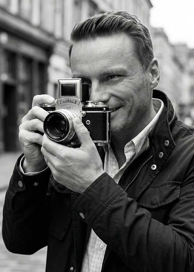 Michael Kormos smiles as he holds a vintage Exakta II SLR camera up to his face, ready to take a photo, while standing outdoors on a New York City street. The image is in black and white.