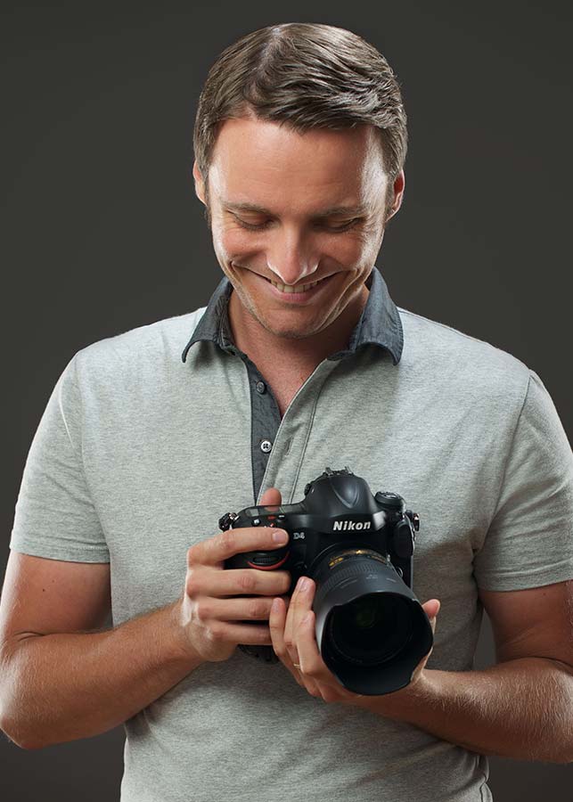 Headshot of Michael Kormos in his NYC photography studio. He is wearing a light gray polo shirt, and smiling while looking down at a Nikon camera that he is holding with both hands against a plain dark background.