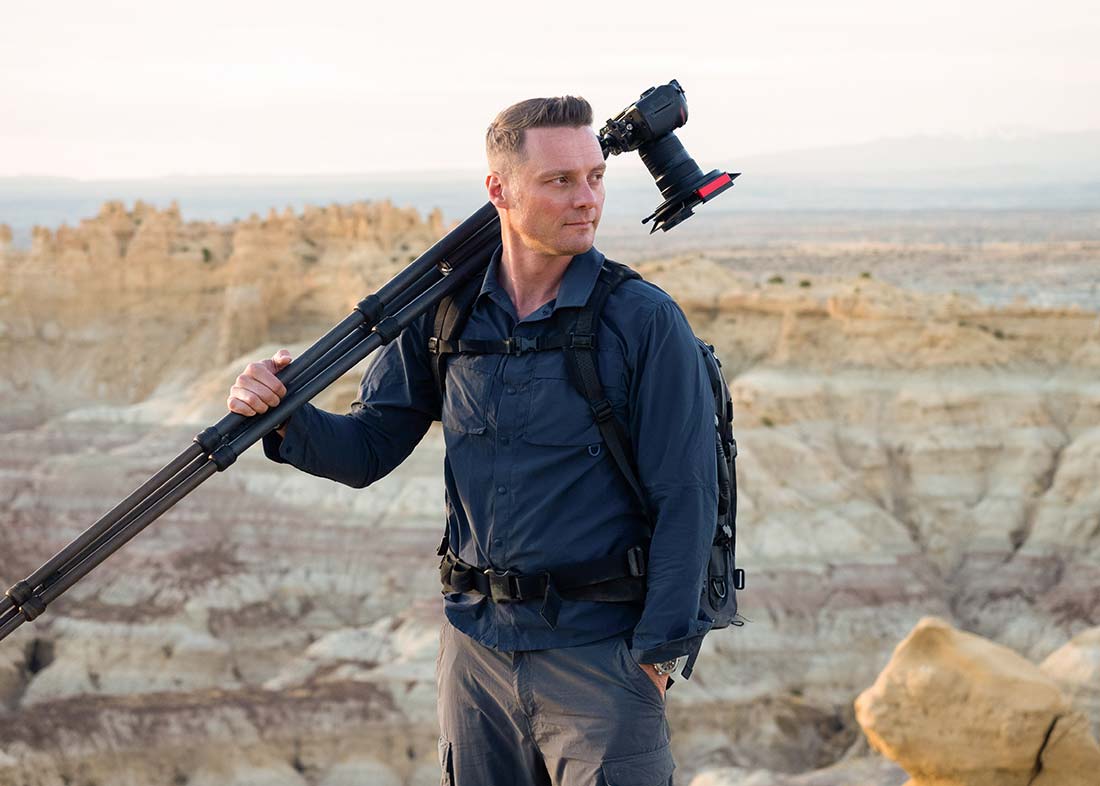 Michael Kormos, wearing a dark shirt and backpack stands outdoors in a rocky, desert landscape, holding a tripod with a camera slung over his shoulder.