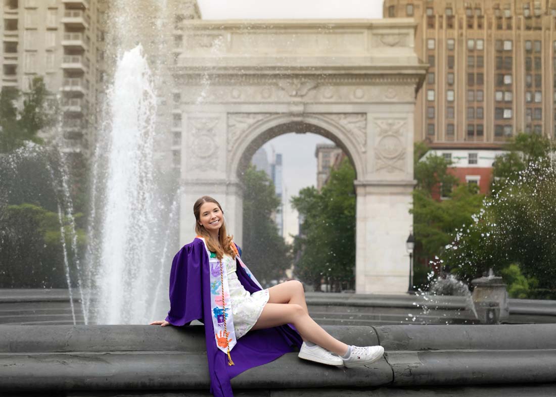 An NYU graduate in a purple gown and white dress smiles on a fountain ledge for her senior portrait, with water spraying behind her in front of the Washington Square Arch at Washington Square Park.