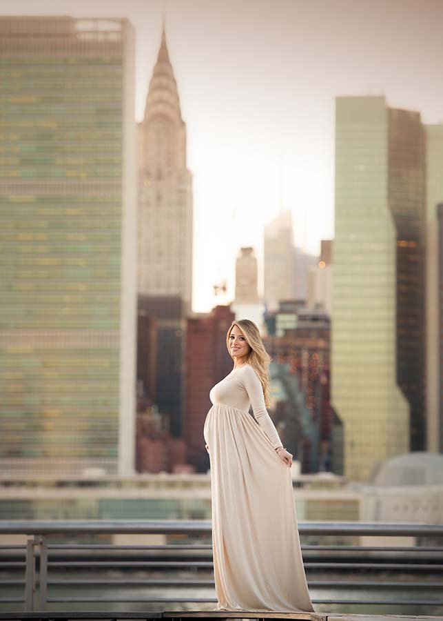 A pregnant woman in a long, flowing cream dress stands on a rooftop in Long Island City NY, smiling, with tall city skyscrapers blurred in the background during daylight—a stunning pregnancy photoshoot.