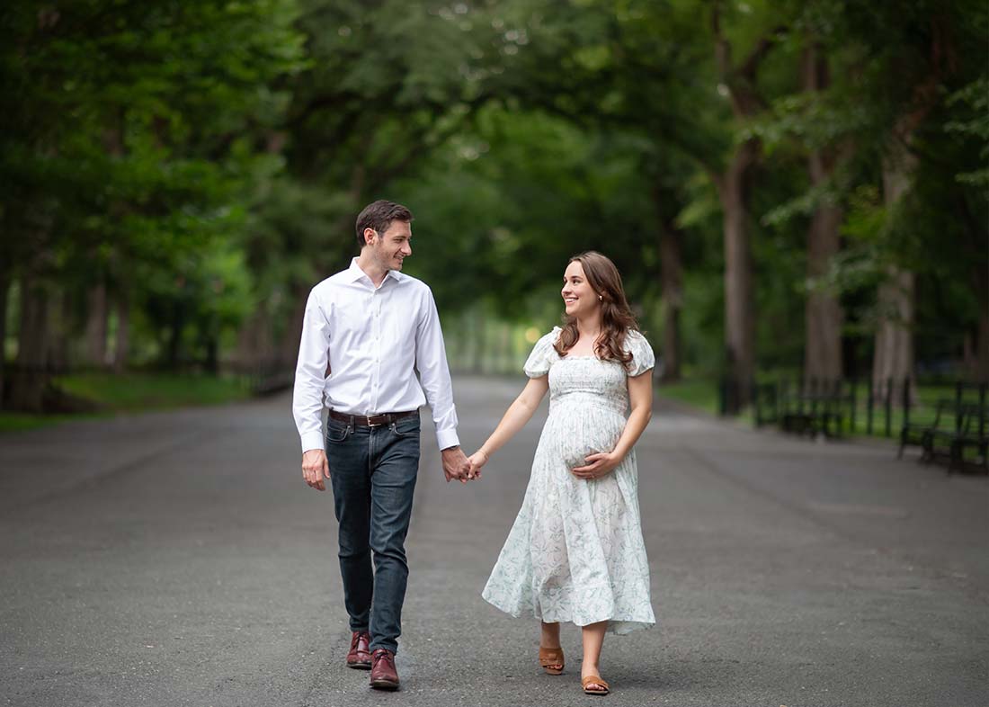 A couple walks hand in hand down a tree-lined path near Bethesda Terrace Central Park. The woman, visibly pregnant in a light floral dress, smiles at the man beside her. The peaceful, green setting adds to their joy.