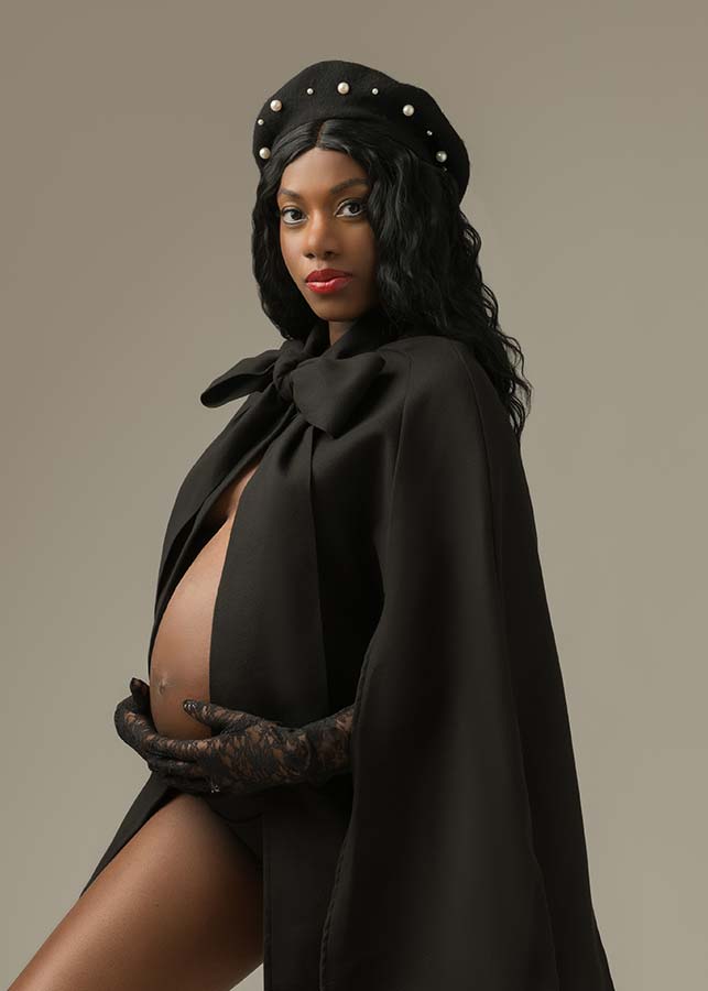 A pregnant woman poses confidently in a fine art pregnancy photoshoot in Manhattan, wearing a black cape with a large bow, a pearl-studded beret, and lace gloves as she cradles her bare baby bump against a neutral background.