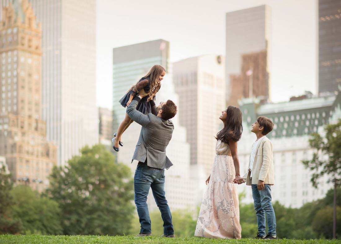A father lifts his young daughter into the air while his wife and their son stand beside them, holding hands, during a family photoshoot in Central Park, with the Plaza Hotel and skyscrapers in the background.