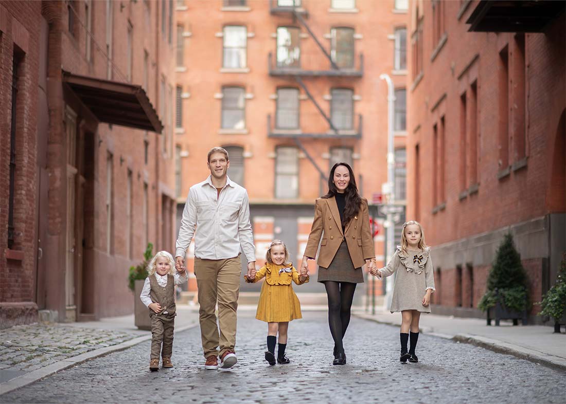 A lifestyle family portrait captures a family of five—two adults and three young children—walking hand-in-hand down a cobblestone street in Tribeca, NYC, all dressed in coordinated fall outfits of brown, beige, and yellow.
