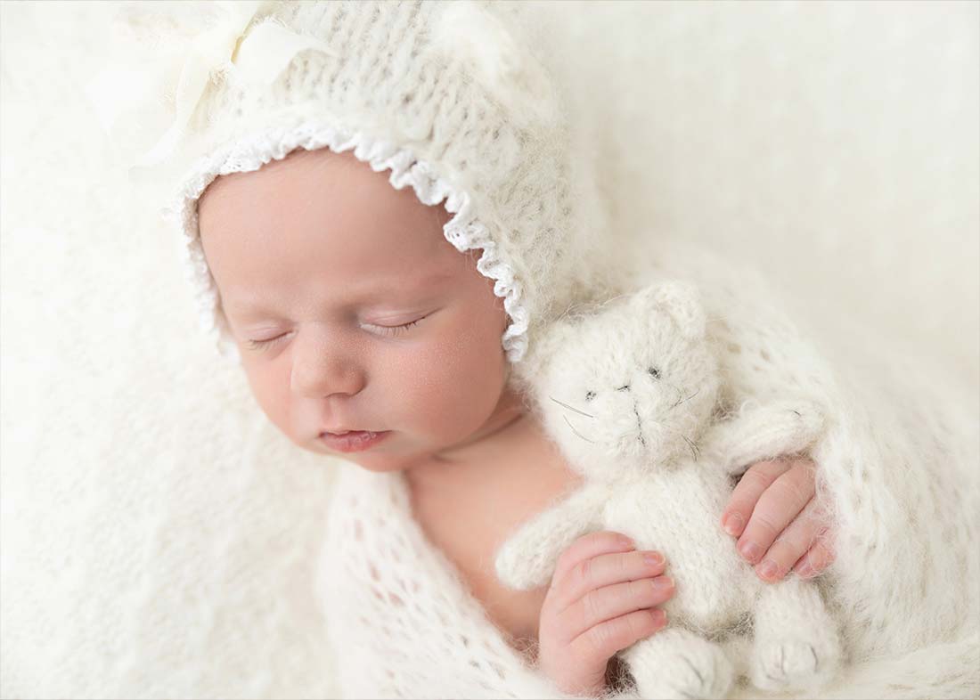 A newborn baby sleeps peacefully, wrapped in a soft white knitted hat and blanket, holding a tiny teddy bear—capturing a cozy and serene moment in this newborn portrait at an NYC photo studio.