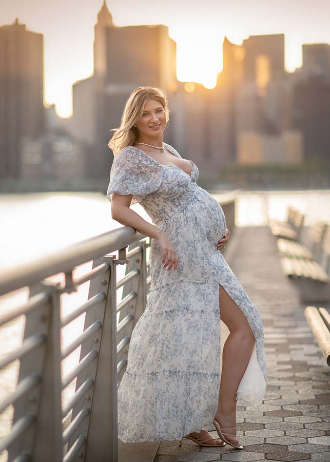 A pregnant woman in a light floral dress stands by a waterfront railing at sunset, smiling and holding her belly during a lifestyle pregnancy photoshoot in Long Island City, with the city skyline in the background.
