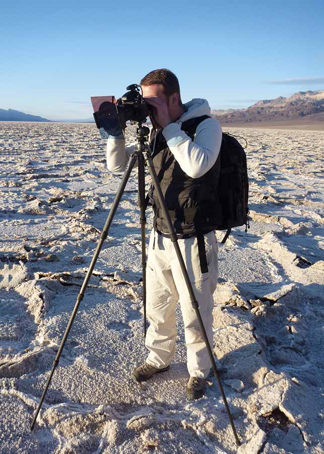 Michael Kormos behind the scenes in Death Valley National Park: He is wearing outdoor gear and a backpack uses a camera on a tripod to take photos on a cracked, salt-covered landscape under a clear sky.