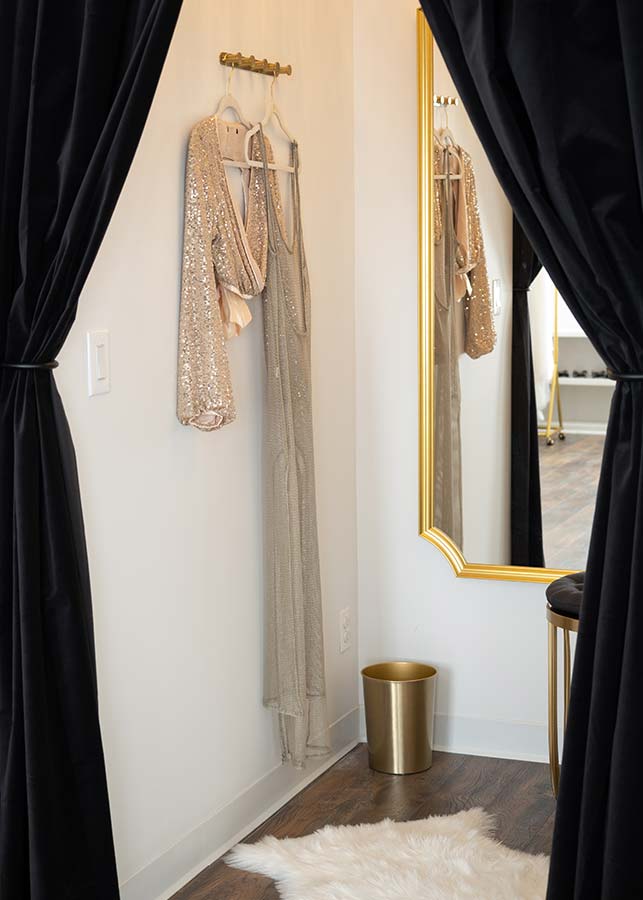 A stylish changing room at the MKP photo studio in NYC features black curtains, a gold-framed mirror, a faux fur rug, sparkly dresses on the wall, a gold trash can, and wooden floors.
