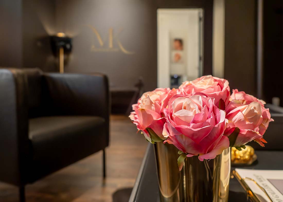 A close-up of a bouquet of pink roses in a gold vase on a table, set in the reception area of Michael Kormos Photography in New York, with a modern, dimly lit lounge and blurred logo visible in the background.