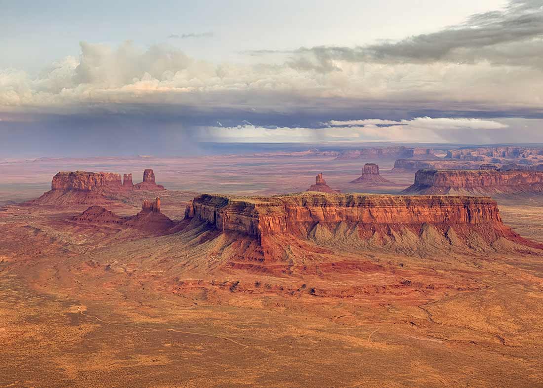 A vast desert landscape featuring dramatic red rock mesas and buttes under a cloudy sky in Monument Valley, with sunlight highlighting the rugged terrain.