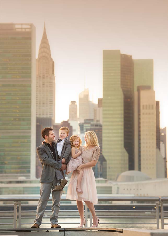 A picturesque family portrait unfolds at sunset as a family of four stands on a rooftop, the Chrysler Building and NYC skyscrapers aglow in the background, parents smiling warmly at their young children held close in their arms.