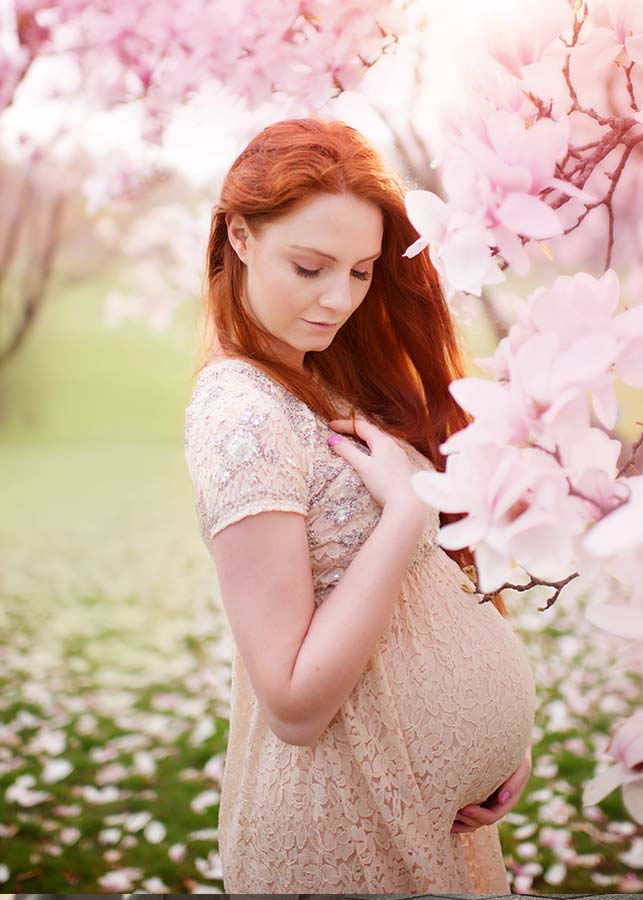 A pregnant woman with long red hair in a light lace dress stands outdoors among blooming cherry blossoms in Central Park, gently touching her belly and looking down softly—a serene moment captured by a skilled maternity photographer.