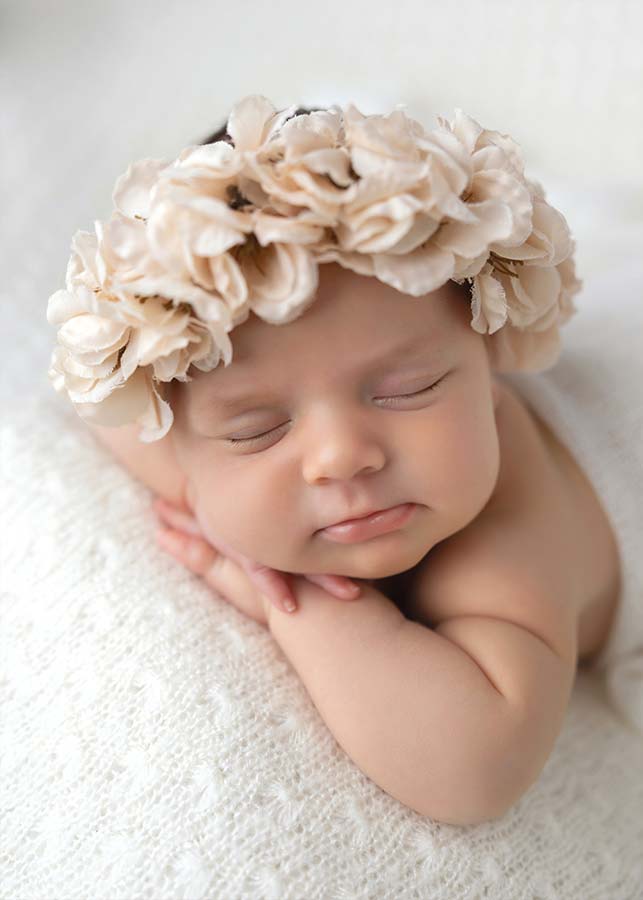 A newborn baby sleeps peacefully on a soft white blanket during a TriBeCa in-home newborn photoshoot, resting their head on their hands. The baby wears a cream-colored floral headband, adding a delicate touch to the serene scene.