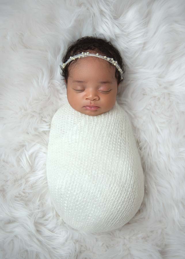 A newborn baby with closed eyes is swaddled in a white blanket, lying on a soft, furry surface, wearing a delicate headband with tiny pearls—perfect for a newborn baby photo session on the Upper West Side.