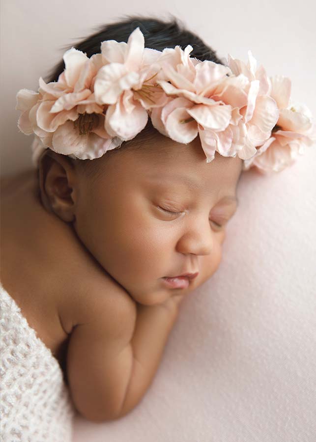 A newborn baby sleeps peacefully on a soft pink blanket, wearing a large light pink flower headband and a white knit wrap—capturing the tender moment as only a lifestyle baby photographer in Manhattan can.